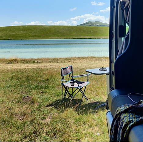 vue depuis un van aménagé au bord d'un lac avec une table et chaise en extérieur
